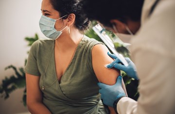 Woman in green short sleeved top being given a vaccination in her left arm by a medical professional