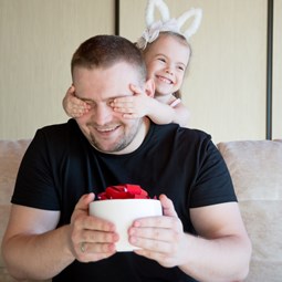 man holding a surprise gift, young child with hands over his eyes